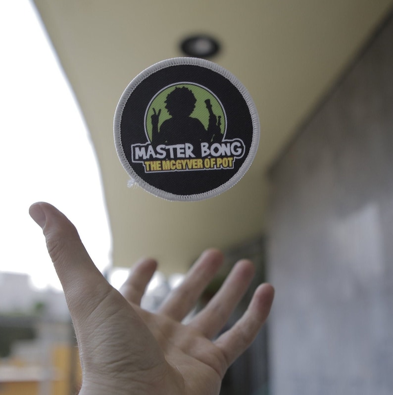 Peut inclure: Un patch rond noir et blanc avec une silhouette verte d'une personne tenant un bang. Le texte sur le patch est "Master Bong The McGyver of Pot".