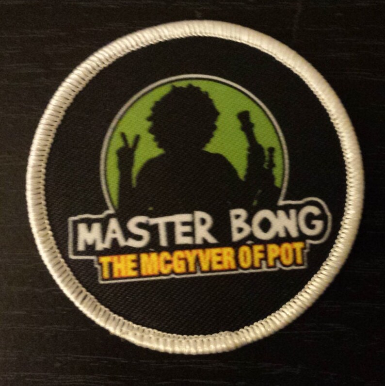 Peut inclure: Un &eacute;cusson brod&eacute; noir et blanc avec un fond vert. L'&eacute;cusson repr&eacute;sente une silhouette d'une personne tenant un signe de paix et un bang. Le texte "Master Bong The MacGyver of Pot" est &eacute;crit en jaune sur l'&eacute;cusson.