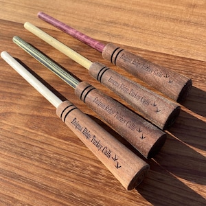 May include: Four wooden turkey calls with the text "Enigma Ridge Turkey Calls" engraved on each. The calls are different colors and have a wood grain pattern.