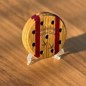 May include: A wooden turkey call with a circular design. The call features a light brown wood grain with two vertical burgundy stripes and eight black holes. The text "Enigma Ridge Turkey Calls" is visible. The call is displayed on a clear stand.