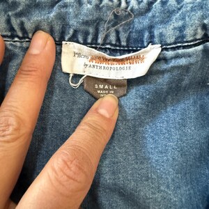 Pu&ograve; includere: Primo piano di una camicia di jeans blu con un'etichetta che recita "Pilcro by Anthropologie". L'etichetta indica anche la taglia "SMALL" e "MADE IN IND". La camicia ha cuciture visibili e un colletto button-down.