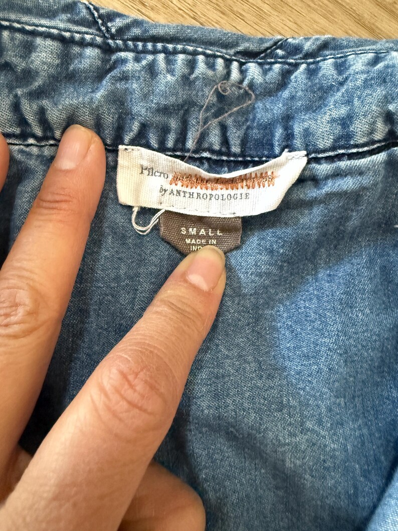 Pu&ograve; includere: Primo piano di una camicia di jeans blu con un'etichetta che recita "Pilcro by Anthropologie". L'etichetta indica anche la taglia "SMALL" e "MADE IN IND". La camicia ha cuciture visibili e un colletto button-down.