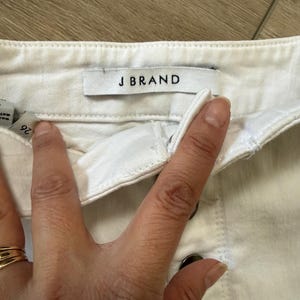 Pu&ograve; includere: Primo piano di pantaloncini di jeans bianchi con l'etichetta del marchio "J BRAND" visibile. I pantaloncini hanno una chiusura con bottoni e un'etichetta della taglia che mostra "26". Il tessuto sembra essere una robusta miscela di cotone.