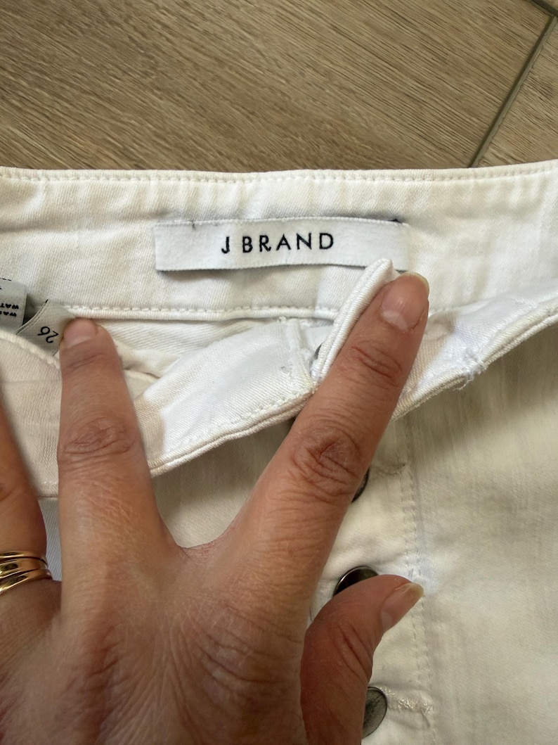 Pu&ograve; includere: Primo piano di pantaloncini di jeans bianchi con l'etichetta del marchio "J BRAND" visibile. I pantaloncini hanno una chiusura con bottoni e un'etichetta della taglia che mostra "26". Il tessuto sembra essere una robusta miscela di cotone.