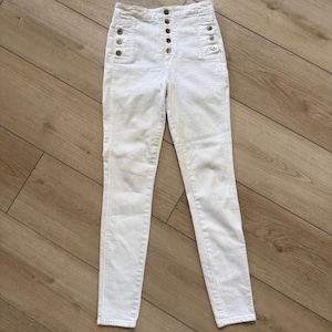 Pu&ograve; includere: Jeans bianchi a vita alta con patta con bottoni e bottoni decorativi sulle tasche. I jeans hanno una vestibilit&agrave; slim e sono realizzati in materiale denim di colore chiaro. I pantaloni sono appoggiati su una superficie di legno.