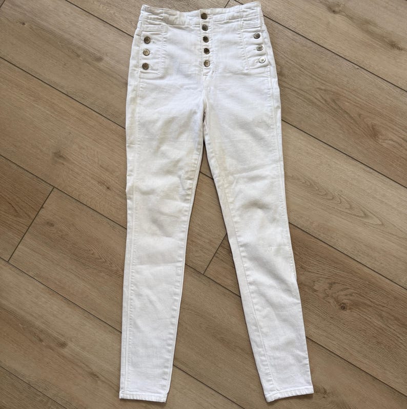Pu&ograve; includere: Jeans bianchi a vita alta con patta con bottoni e bottoni decorativi sulle tasche. I jeans hanno una vestibilit&agrave; slim e sono realizzati in materiale denim di colore chiaro. I pantaloni sono appoggiati su una superficie di legno.