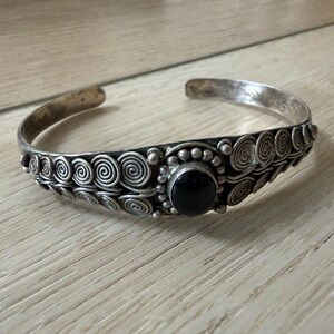 May include: A silver cuff bracelet with a central black stone and spiral design. The bracelet has an open design and is made of a silver-toned metal. The bracelet is approximately 2.5 inches in diameter.