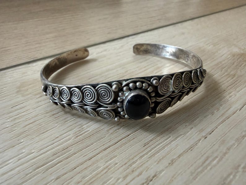 May include: A silver cuff bracelet with a central black stone and spiral design. The bracelet has an open design and is made of a silver-toned metal. The bracelet is approximately 2.5 inches in diameter.