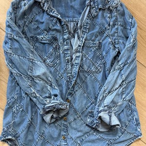 Pu&ograve; includere: Una camicia di jeans azzurra con un esclusivo motivo a rombi. La camicia a maniche lunghe presenta due tasche frontali e una chiusura con bottoni. I polsini sono arrotolati e la camicia &egrave; stesa su una superficie di legno.