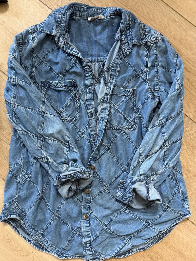 Pu&ograve; includere: Una camicia di jeans azzurra con un esclusivo motivo a rombi. La camicia a maniche lunghe presenta due tasche frontali e una chiusura con bottoni. I polsini sono arrotolati e la camicia &egrave; stesa su una superficie di legno.