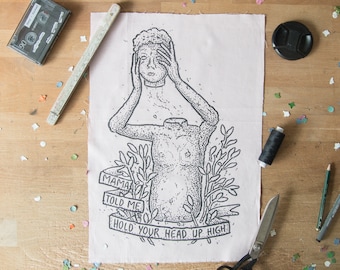 May include: A rectangular piece of light pink fabric featuring a black ink illustration of a headless torso holding its own head. The design includes leafy branches and a banner with the text MAMA TOLD ME HOLD YOUR HEAD UP HIGH.