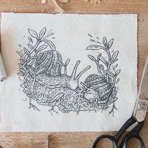 May include: A rectangular piece of off-white fabric featuring a black ink illustration of two snails crawling on a mossy patch surrounded by leafy stems. To the left are two spools of thread, one black and one white, and to the right are metal scissors.