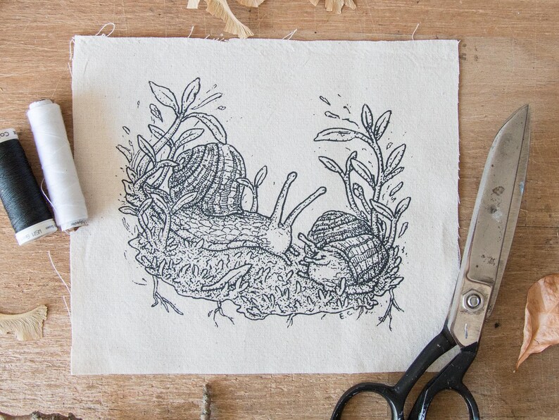 May include: A rectangular piece of off-white fabric featuring a black ink illustration of two snails crawling on a mossy patch surrounded by leafy stems. To the left are two spools of thread, one black and one white, and to the right are metal scissors.