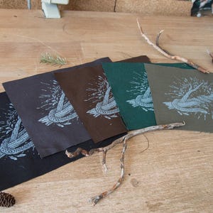 May include: Four square fabric patches in black, dark brown, dark green, and olive green, each featuring a white screen-printed graphic of a knotted rope entwined with leafy branches. The patches are arranged in a fan shape on a wooden surface.