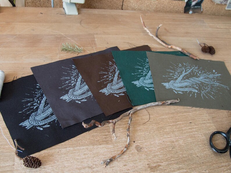 May include: Four square fabric patches in black, dark brown, dark green, and olive green, each featuring a white screen-printed graphic of a knotted rope entwined with leafy branches. The patches are arranged in a fan shape on a wooden surface.