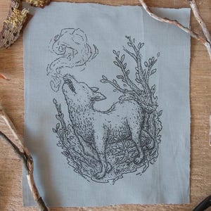 Könnte beinhalten: Hellblaues Stoffquadrat mit einer schwarzen Illustration eines heulenden Wolfes, umgeben von Blättern. Der Wolf blickt nach oben, mit einem Rauchwölkchen aus dem Maul. Zweige und Blätter umgeben das Kunstwerk.