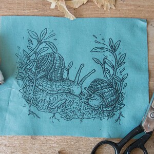 May include: A rectangular piece of teal fabric featuring a black ink illustration of two snails crawling on mossy ground surrounded by leafy stems. The initials E.C. are printed in the bottom right corner of the illustration on the fabric.