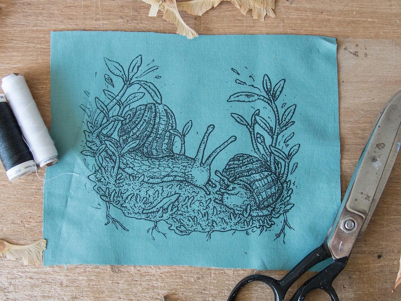 May include: A rectangular piece of teal fabric featuring a black ink illustration of two snails crawling on mossy ground surrounded by leafy stems. The initials E.C. are printed in the bottom right corner of the illustration on the fabric.