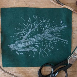 May include: A rectangular piece of dark green fabric featuring a white screen-printed illustration of a snake coiled around a tree branch. The design includes radiating lines and small leaves surrounding the central snake and branch motif.