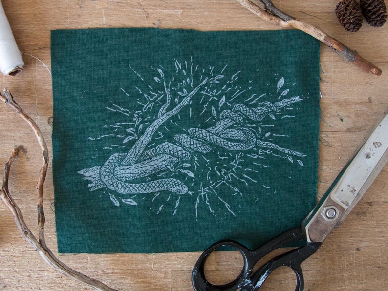 May include: A rectangular piece of dark green fabric featuring a white screen-printed illustration of a snake coiled around a tree branch. The design includes radiating lines and small leaves surrounding the central snake and branch motif.