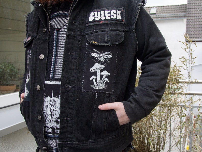 May include: A black denim vest featuring a rectangular patch with a white graphic of mushrooms and a winged insect. The vest includes a rectangular patch with the text Kylesa and another patch with a detailed black and white illustration.