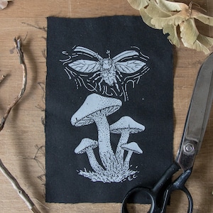 May include: A rectangular black fabric patch featuring a white screen-printed design of a cluster of mushrooms at the bottom and a winged insect at the top. The print includes fine stippling details and the initials E.C. in the bottom right corner.