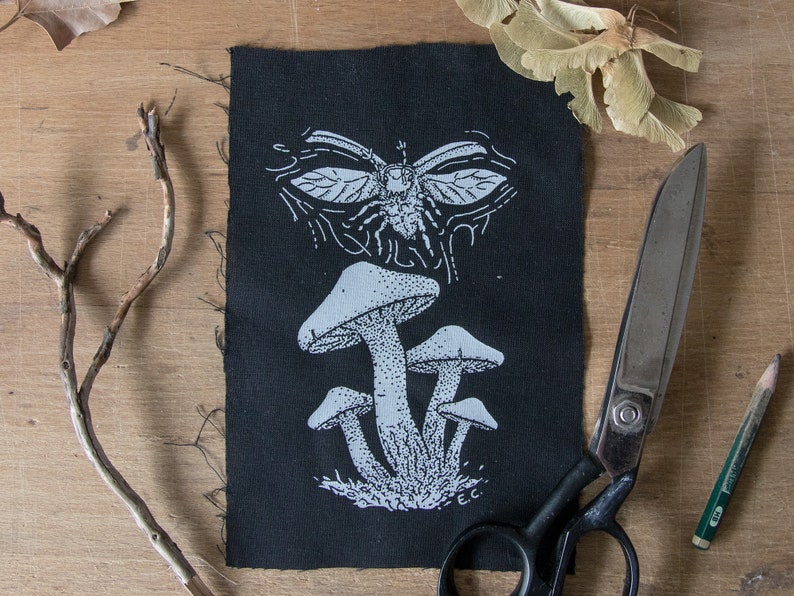 May include: A rectangular black fabric patch featuring a white screen-printed design of a cluster of mushrooms at the bottom and a winged insect at the top. The print includes fine stippling details and the initials E.C. in the bottom right corner.