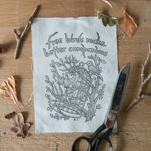 May include: A black and white illustration of a bird in a cage with the text "Free birds make better companions" printed on a white fabric background.