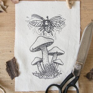 May include: A rectangular piece of off-white fabric featuring a black ink illustration of a cluster of four mushrooms with a flying beetle positioned above them. The fabric has raw, frayed edges and is displayed alongside a pair of metal scissors.