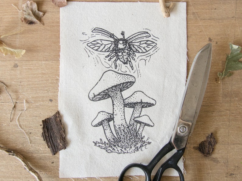 May include: A rectangular piece of off-white fabric featuring a black ink illustration of a cluster of four mushrooms with a flying beetle positioned above them. The fabric has raw, frayed edges and is displayed alongside a pair of metal scissors.