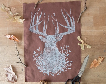 May include: A rectangular brown fabric panel featuring a light blue screen-printed illustration of a deer head with large antlers, surrounded by foliage at the base. The design includes intricate stippled texture and the initials E.L. in the corner.
