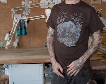 May include: A brown t-shirt with a white graphic print of a group of animals in a forest setting. The person wearing the shirt has several tattoos on their arms.