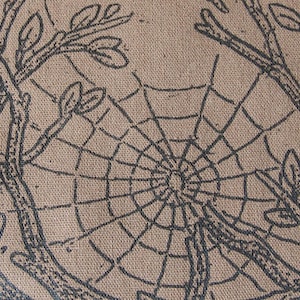 Op de afbeelding: Een beige stoffen oppervlak met een donkerbruine zeefdrukillustratie van een spinnenweb gecentreerd tussen twee boomtakken met bladeren. De textuur van het geweven materiaal is duidelijk zichtbaar in het gehele gedrukte ontwerp.