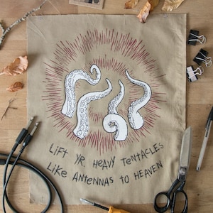Op de afbeelding: Een bruin textiel met een handgetekende illustratie van witte tentakels die uitstralen vanuit een centraal punt, met de tekst "Lift Yr. Heavy Tentacles Like Antennas to Heaven" in het zwart. Verschillende objecten omringen het textiel.