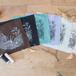 May include: A set of six square fabric patches in brown, black, teal, lavender, white, and cream, each featuring a white or black screen-printed illustration of two snails among foliage. Two small spools of black and white sewing thread are included.