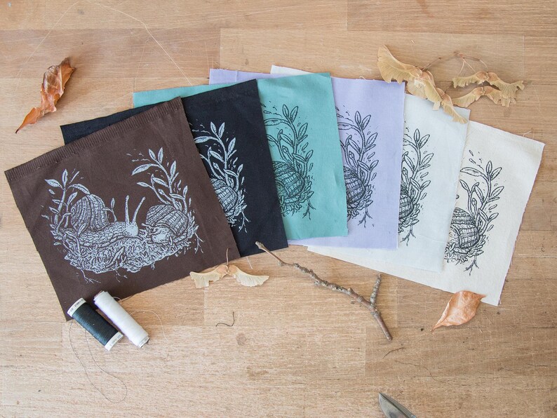 May include: A set of six square fabric patches in brown, black, teal, lavender, white, and cream, each featuring a white or black screen-printed illustration of two snails among foliage. Two small spools of black and white sewing thread are included.