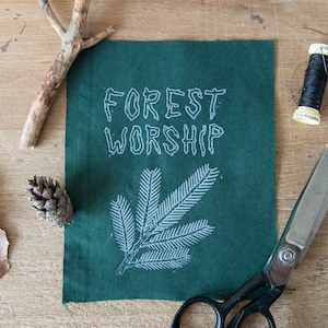 Puede incluir: Un parche de tela verde con bordado blanco que dice "FOREST WORSHIP". El parche presenta un diseño estilizado de rama de pino blanco.
