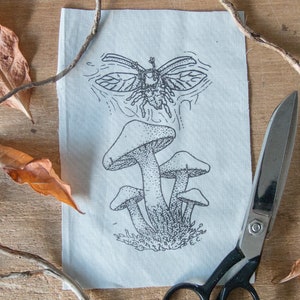 May include: A rectangular piece of off-white fabric featuring a black ink illustration of a cluster of mushrooms with a flying beetle above them. The fabric is positioned next to a pair of black metal scissors and several dried brown leaves.