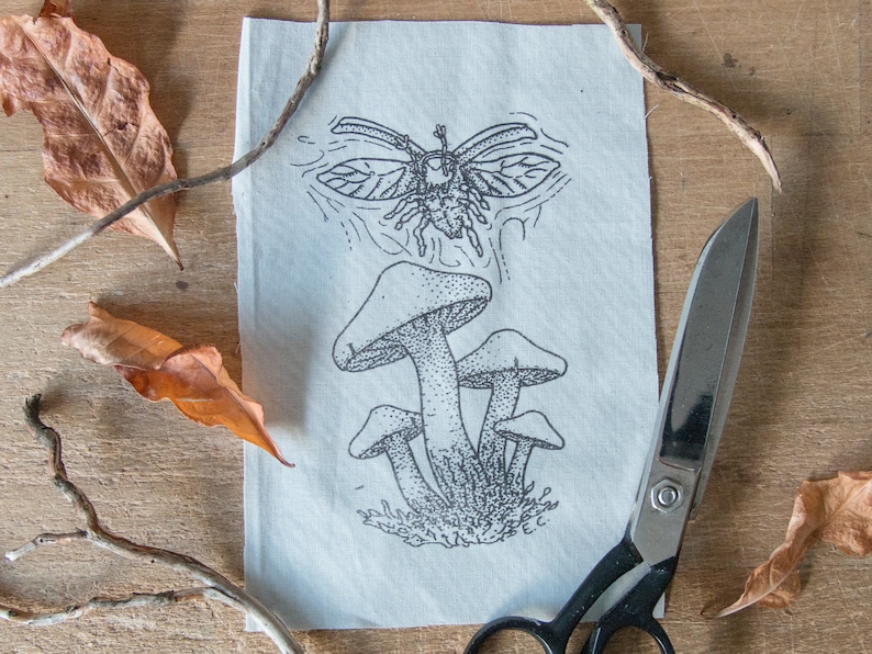 May include: A rectangular piece of off-white fabric featuring a black ink illustration of a cluster of mushrooms with a flying beetle above them. The fabric is positioned next to a pair of black metal scissors and several dried brown leaves.