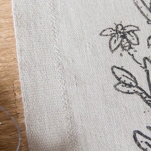Op de afbeelding: Een stuk gebroken wit, gestructureerd textiel met een zwarte inktillustratie van een tak met knoppen en een klein insect. De stof heeft een zichtbare geweven structuur en een rechte, afgewerkte rand die verticaal langs de linkerkant van de print loopt.