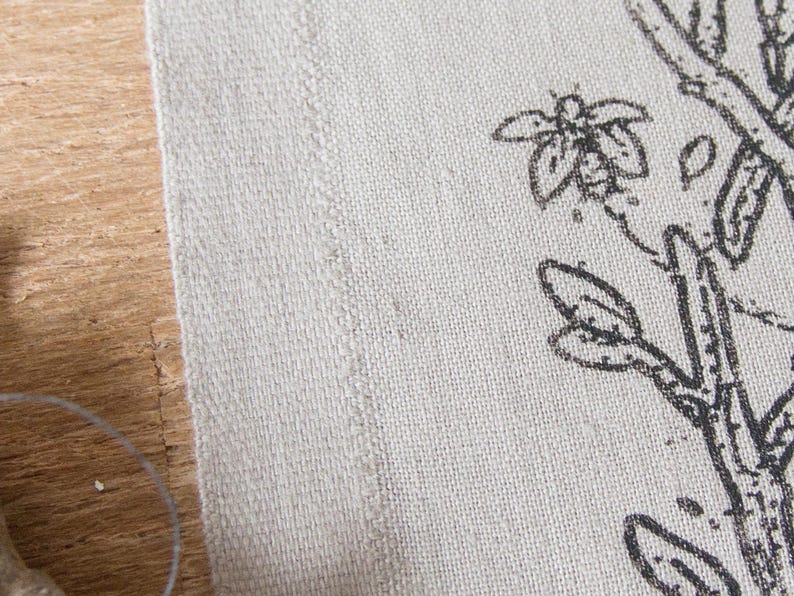 Op de afbeelding: Een stuk gebroken wit, gestructureerd textiel met een zwarte inktillustratie van een tak met knoppen en een klein insect. De stof heeft een zichtbare geweven structuur en een rechte, afgewerkte rand die verticaal langs de linkerkant van de print loopt.