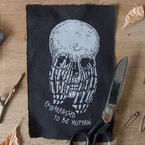 May include: Black and white embroidered patch with a skull design and the text "Embarrassed to be Human".