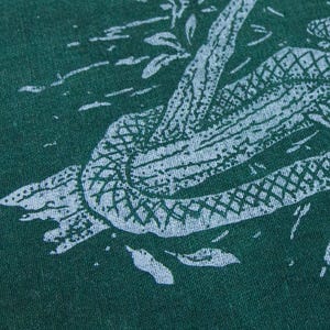 May include: A dark green fabric featuring a light blue screen-printed graphic of a snake coiled around a textured tree branch. The snake has a visible scale pattern, and the design includes small light blue marks representing leaves or debris.