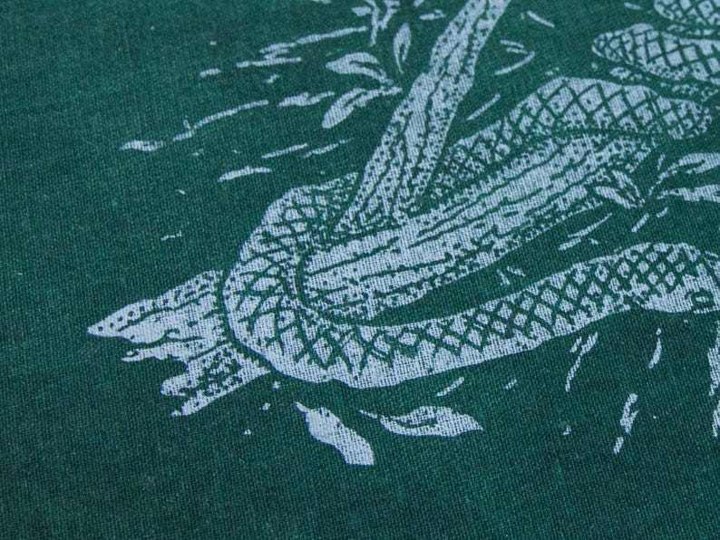 May include: A dark green fabric featuring a light blue screen-printed graphic of a snake coiled around a textured tree branch. The snake has a visible scale pattern, and the design includes small light blue marks representing leaves or debris.