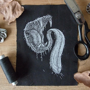 Può includere: Toppa in tessuto nero con una grafica bianca di una creatura ringhiante con denti affilati e una lunga lingua sottile. La lingua della creatura è arrotolata a forma di spirale.