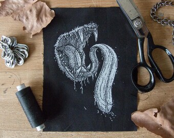 May include: Black fabric patch with a white graphic of a snarling, toothy creature with a long, thin tongue. The creature's tongue is curled into a spiral shape.