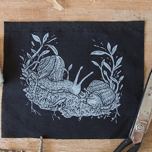 May include: A rectangular black fabric patch featuring a light blue screen-printed illustration of two snails resting on a patch of moss surrounded by leafy stems. The design includes intricate line work detailing the shells and the snail bodies.