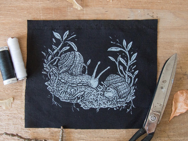 May include: A rectangular black fabric patch featuring a light blue screen-printed illustration of two snails resting on a patch of moss surrounded by leafy stems. The design includes intricate line work detailing the shells and the snail bodies.