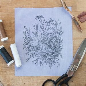 May include: A light purple fabric square featuring a black line art illustration of a snail surrounded by foliage. The fabric is arranged with three spools of thread in brown, black, and white, a pair of metal scissors, and two small twigs.