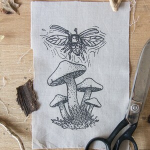May include: A rectangular piece of light beige fabric featuring a black ink illustration of a cluster of mushrooms with a beetle flying above them. The design uses stippling and line work to create texture. A pair of metal scissors is positioned nearby.